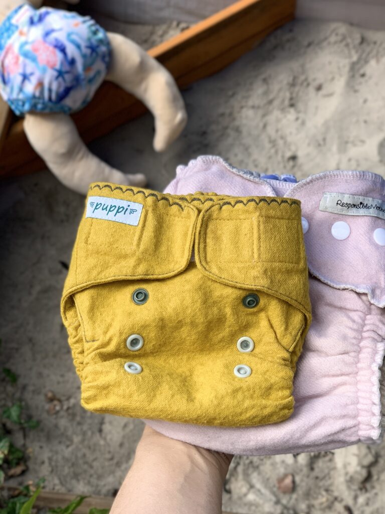 Eine Hand hält eine gelbe Puppi-Wollüberhose und eine altrosa Wollüberhose von Responsible Mother. Im Hintergrund ein mit Sand befüllter Sandkastn. 